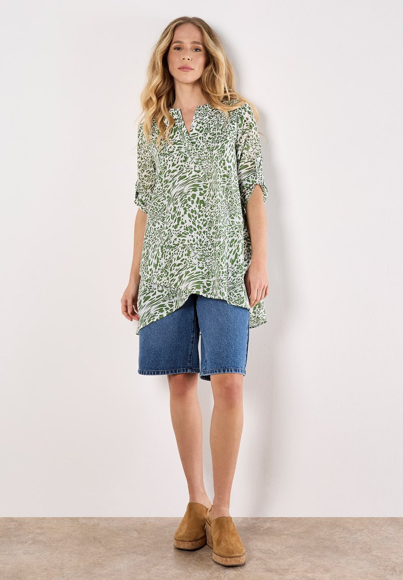 Camisa larga verde y blanca con un patrón y escote en V, combinada con pantalones cortos de mezclilla azul y zuecos marrones, con un fondo neutro.