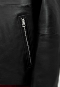 Schwarze Lederjacke mit einer silbernen Reißverschlusstasche am linken Ärmel, glatte Textur und klare Linien mit leichter Polsterung an den Schultern.