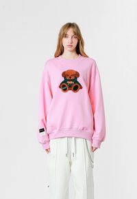 Sudadera rosa con bordado de un osito en marrón y verde. Presenta mangas anchas y un cuello de canalé. Acentuada con etiquetas negras en las mangas.