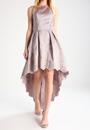 Mujer con vestido lavanda sin mangas de corte asimétrico con detalles de encaje y dobladillo festoneado, combinado con tacones blancos con tiras al tobillo.