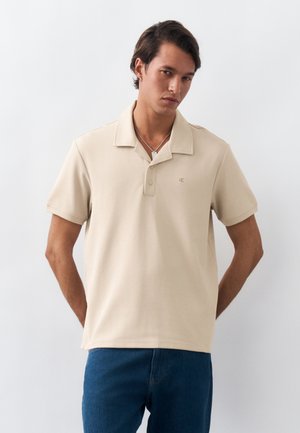 Férfi bézs rövid ujjú galléros pólóban és kék farmerban, kezével a háta mögött áll, egyszínű fehér háttér előtt.