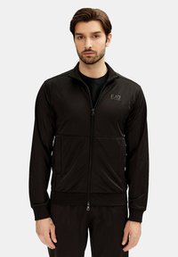 Sort zip-jakke lavet af glat stof, med sidelommer, ribbede manchetter og et lille logo på brystet. Slim fit design.