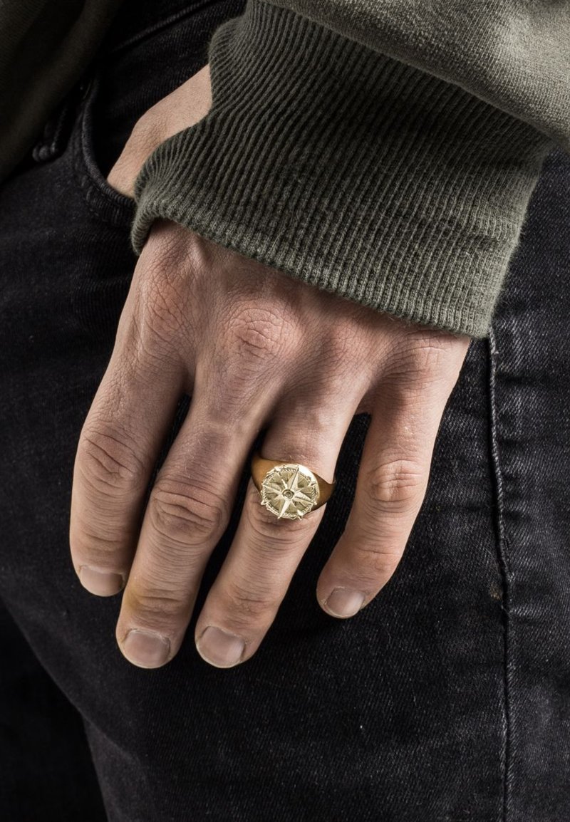 Arany iránytű témájú gyűrű bonyolult részletekkel, egy kézen viselve. A kéz napbarnított bőrszíne denim anyagot fog át, olívazöld ujjal.