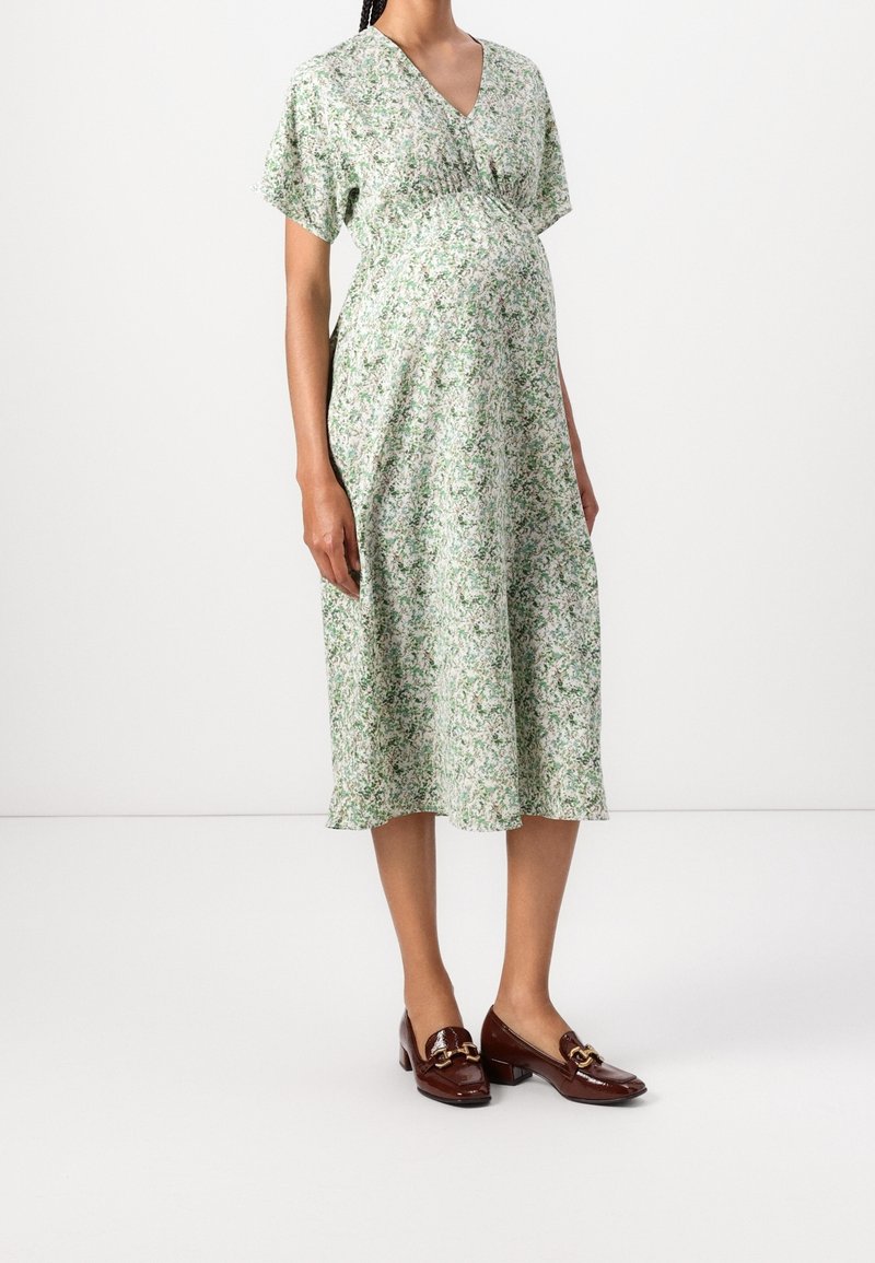 Femme enceinte portant une robe midi fleurie verte et blanche à manches courtes et des mocassins marron à talons bas, debout sur un fond blanc.
