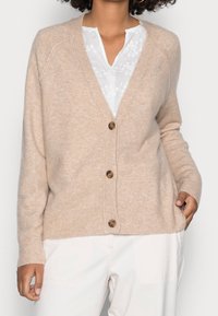 Beige villainen neuletakki V-aukkokoristeella, napit edessä ja resorit hihoissa. Pehmeä materiaali ja rento istuvuus. Päällä valkoinen paita.