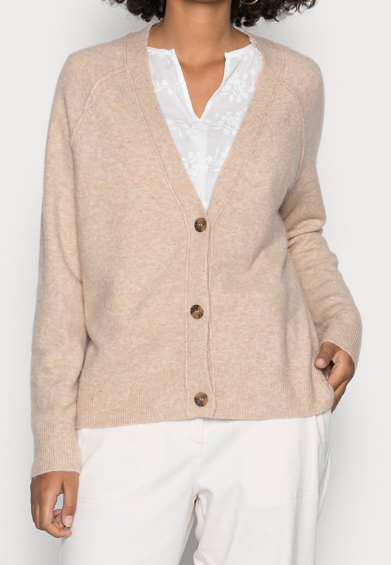 Beige villainen neuletakki V-aukkokoristeella, napit edessä ja resorit hihoissa. Pehmeä materiaali ja rento istuvuus. Päällä valkoinen paita.