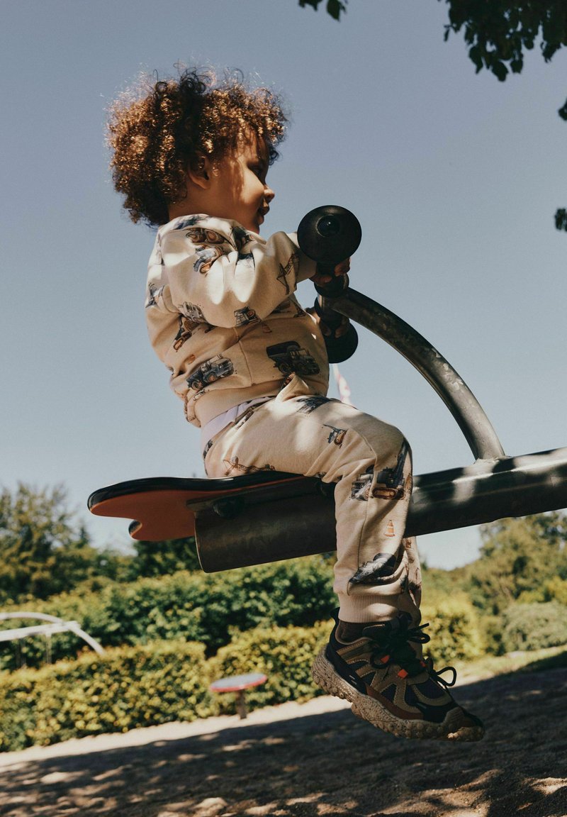 Enfant aux cheveux bouclés portant des vêtements à motifs joue sur une bascule dans une aire de jeux extérieure ensoleillée avec de la verdure en arrière-plan.