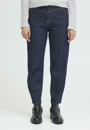 Person iført mørkeblå jeans med lige ben, sorte læderankelstøvler og en hvid langærmet skjorte med sorte striber.