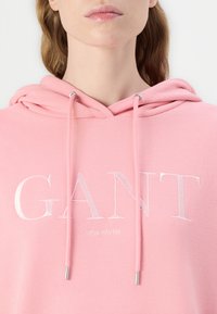 Ljusrosa hoodie i mjukt tyg, med dragsnören med metallspetsar och en vit broderad logotyp "GANT NEW HAVEN" på framsidan.