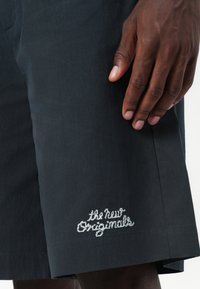 Donkergrijze shorts met een gladde textuur, met wit geborduurde tekst "de nieuwe Originals" nabij de zoom. Close-up van een hand op de stof.