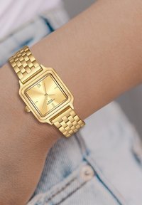 Kuldtooniline ruudukujuline kell, millel on tekstuuriga dial, kahe käsiraami näidikud ja metallvõrksidemed käel, heleda teksakanga taustal.