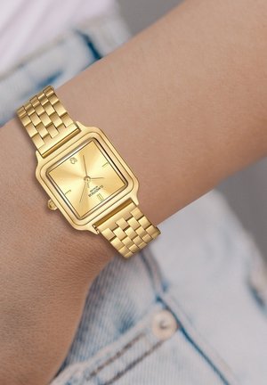 Montre carrée dorée avec un cadran texturé, deux indicateurs d'heure et un bracelet en maille métallique sur un poignet, le tout sur un fond en denim clair.