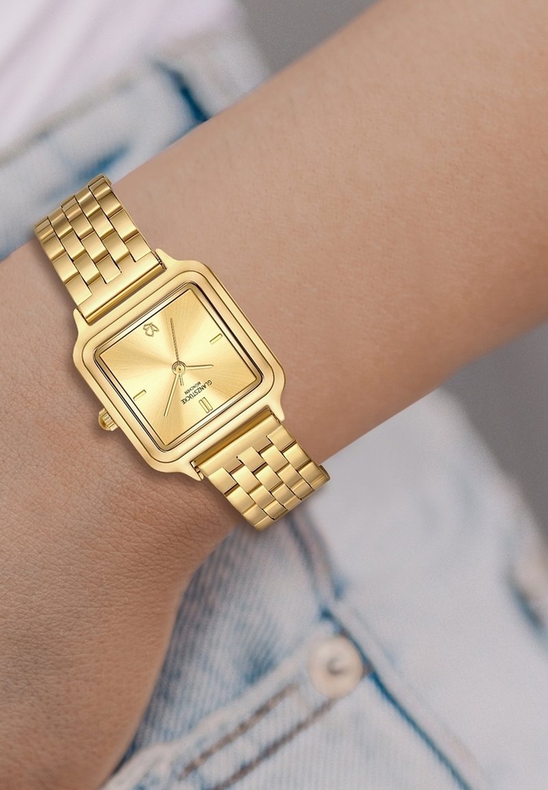 Kuldtooniline ruudukujuline kell, millel on tekstuuriga dial, kahe käsiraami näidikud ja metallvõrksidemed käel, heleda teksakanga taustal.