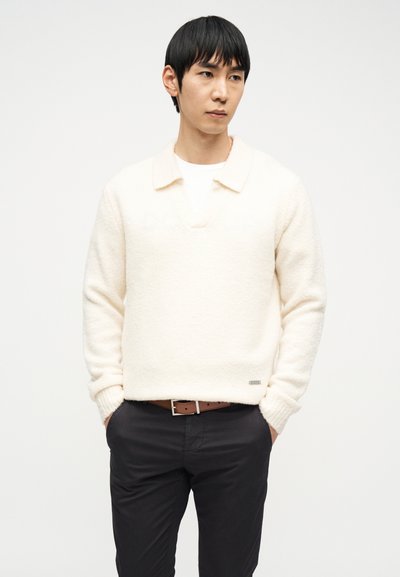 Άνδρας με μαύρα μαλλιά που φοράει κρεμ πουλόβερ με υφή και γιακά, λευκό εσώρουχο, καφέ ζώνη και μαύρο παντελόνι, στέκεται με τα χέρια στις τσέπες.