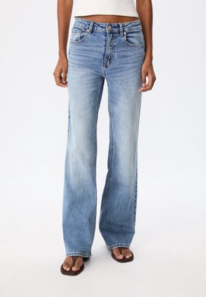 Frau trägt hellblaue High-Waist-Jeans mit weitem Bein, vorderen Taschen, Knopfleiste, weißes, kurz geschnittenes Oberteil und braune Zehenstegsandalen.