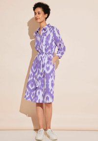 Lila-weißer gemusterter Kleid mit Knopfverschluss vorne, einem Taillengürtel und zwei Seitentaschen. Kombiniert mit weißen Sneakers.