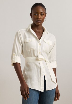 Cămașă albă din in cu mâneci rulabile, nasturi în față, două buzunare la piept și o curea din material legată în talie. Textură netedă și strălucire subtilă.