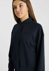 Marineblauwe trackjack gemaakt van glad materiaal, met een hoge kraag, ritssluiting, zijzakken en een geborduurd logo op de borst.