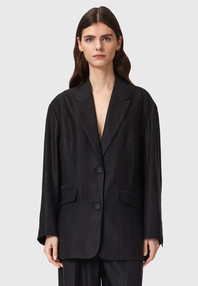 Femme aux longs cheveux bruns portant un blazer noir oversize à deux boutons avec des poches à rabat sur le devant, debout devant un fond gris.