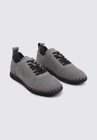 Zapatillas de malla gris con cordones negros, parte superior perforada, suela de goma con detalles texturizados y cinta en el talón para ponerlas y quitarlas fácilmente.