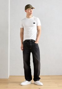 T-shirt bianco a maniche corte con tasca nera, abbinato a jeans dritti di colore blu scuro e sneakers bianche. Un cappellino verde oliva completa l'outfit.