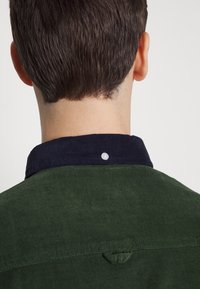 Grön kordflortskjorta med en marinblå krage och en knappdetalj baktill. Har ett texturerat tyg och en ögla vid halsen.