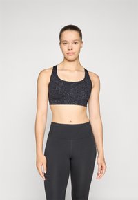Crni sportski grudnjak s suptilnim uzorkom leopard printa, napravljen od rastezljivog materijala, u kombinaciji s visokim crnim tajicama.
