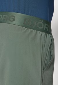 Pantalones cortos verdes con una cintura texturizada que presenta la palabra "BJÖRG" en un verde más oscuro. Bolsillos laterales con un acabado de tela suave.