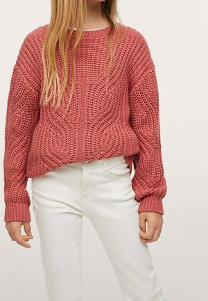 Kobieta ubrana w grubą dzianinową bluzę w kolorze pudrowego różu z długim rękawem i białe spodnie, stojąca na tle jasnego, jednolitego tła.