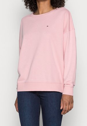 Kvinde iført en lyserød langærmet sweatshirt med rund hals og lille logo på brystet samt mørkeblå jeans.