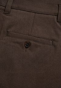 Tissu brun foncé en gros plan montrant une poche arrière de pantalon avec un seul bouton noir et des passants de ceinture.