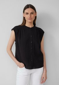 Blouse noire à manches courtes en tissu léger et froissé, dotée d'un bouton sur le devant et d'une coupe décontractée. Un pantalon blanc est porté en dessous.