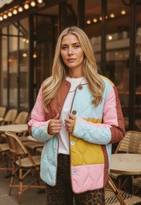 Blondínka v farebnej prešívanej bunde stojí v vonkajšej kaviarni s prútenými stoličkami a stolmi v pozadí.