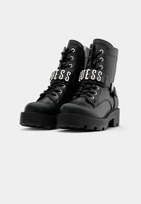 Botas de combate negras con suela de goma gruesa, material sintético brillante, diseño de cordones y una correa de marca plateada alrededor del tobillo.