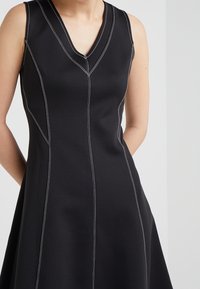 Vestito nero senza maniche con scollatura a V, caratterizzato da cuciture a contrasto bianche lungo le cuciture verticali e un'orlo svasato. Tessuto liscio e testurizzato.