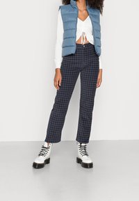 Světle modrá vatovaná vesta, bílý top se stahovací šňůrkou, námořnicky modré kostkované kalhoty a bílé boty na šněrování s černými podrážkami. Jednolitá textura, střih na míru.