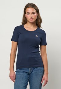 Ung kvinde med langt brunt hår iført en tætsiddende mørkeblå kortærmet skjorte og blå jeans, stående mod en ensartet lys baggrund.