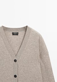 Beige bomullskardigan med djup V-ringning, två svarta knappar och ribbade ärmslut. Mjuk textur med en avslappnad och ledig passform.