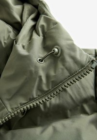 Jachetă puffer de culoare verde măslinie, cu textură moale, matlasată, închidere cu fermoar metalic, cordon cu ochi și finisaj de țesătură netedă.