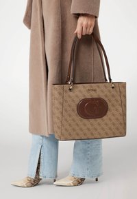 Brun mønstret shoppingtaske med et fremtrædende logo, dobbelte håndtag og guldfarvet hardware. Kombineret med lyseblå jeans og slangeskindsprintede hæle.