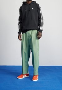 Musta huppari, jossa valkoiset raidat hihoissa, yhdistettynä löysiin vihreisiin housuihin ja oransseihin lenkkareihin, joissa valkoiset yksityiskohdat.