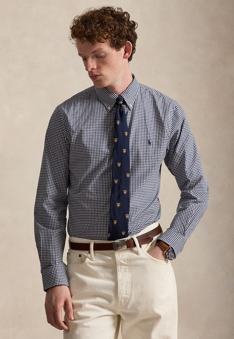 Homme portant une chemise à carreaux bleue à boutons, une cravate marine avec des blasons, un pantalon crème, une ceinture en cuir marron et une montre-bracelet, debout avec la main dans la poche.