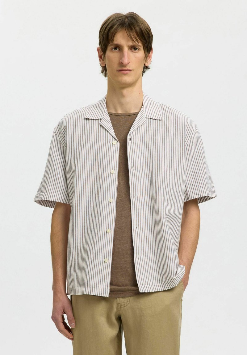 Ung mand iført en kortærmet beige og hvid skjorte med lodrette striber over en brun T-shirt og khakibukser, stående mod en ensfarvet baggrund.