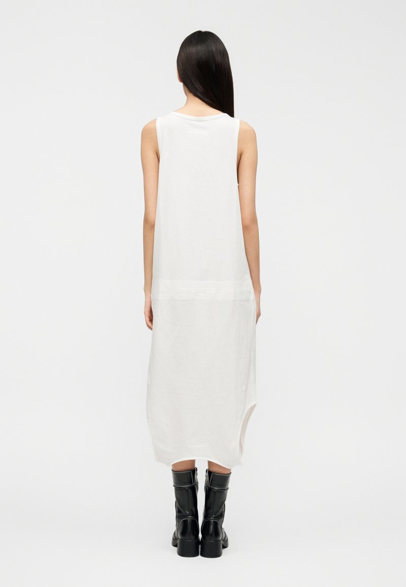 Robe blanche sans manches en tissu doux, avec un col arrondi et un ourlet asymétrique. Portée avec des bottes noires montantes jusqu'aux genoux.