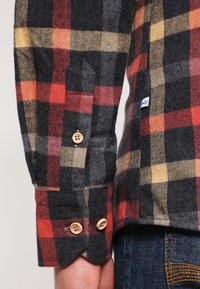 Flanelska košulja s crno, crvenom i žutom kariranom uzorkom. Ima dugme na manžetama, herringbone teksturu i oznaku s logotipom na rukavu.