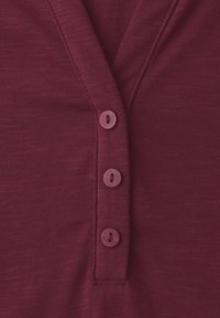 Nærbillede af vinrødt stof med tre matchende runde knapper på en knaplist, der fremhæver den teksturerede strik og syningsdetaljer.