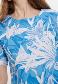 Sininen lyhythihainen paita, jossa on valkoinen trooppinen lehtikuosi. Kangas näyttää kevyeltä ja tekstuuriltaan rennolta, ja se istuu väljästi.