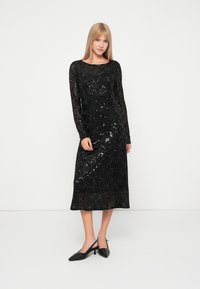 Vestido preto de lantejoulas com mangas longas e decote barco, apresentando uma silhueta ajustada e comprimento até ao joelho, combinado com sapatos de salto tipo slingback pretos.