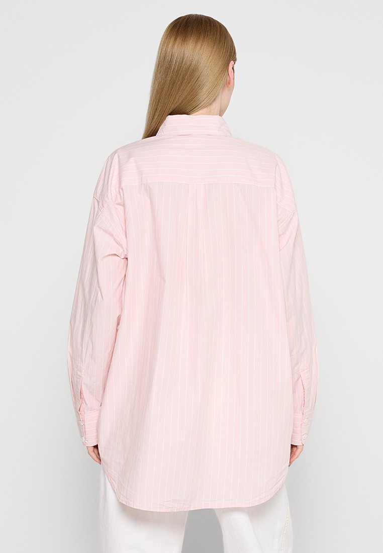 Chemise button-down rose clair avec des rayures blanches verticales, à manches longues et à coupe décontractée. Dotée d'un col pointu et d'un détail de empiècement au dos.