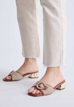 Pies con sandalias beige de tacón abierto con hebillas decorativas combinadas con pantalones de color claro con dobladillo deshilachado sobre fondo blanco.